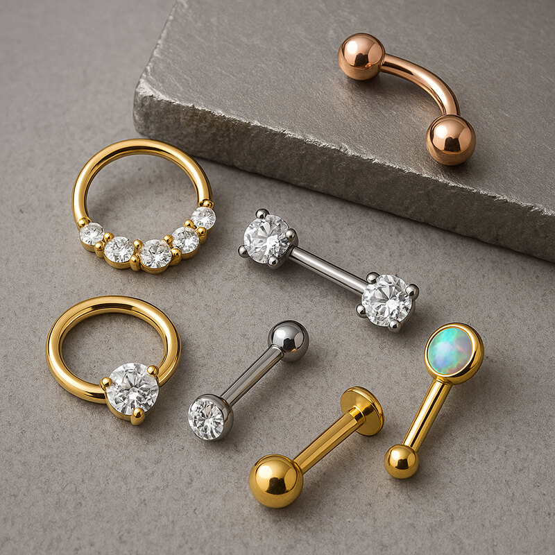 Assorted body jewellery on a grey stone surface, including gold and silver barbells, rings with clear gemstones, and a gold labret with an opal setting.